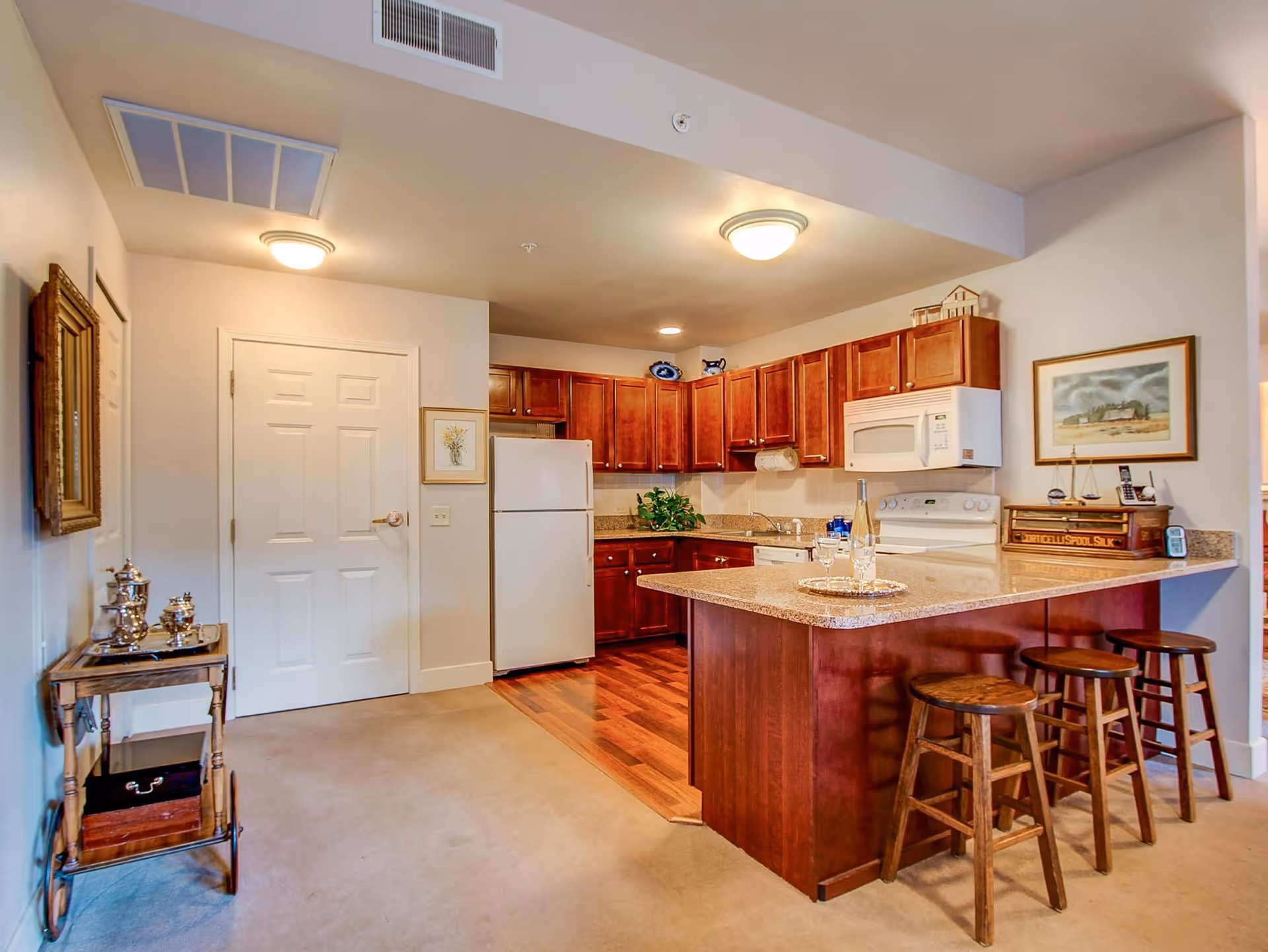 Interior view of a kitchen area in St. Andrew's Village featuring wooden cabinets, a white refrigerator, a white microwave, and a stove. There is a kitchen island with a granite countertop and three wooden bar stools. The floor transitions from carpet to wood in the kitchen area. A small table with silver tea set is on the left side near a closed white door. The walls are decorated with framed artwork.