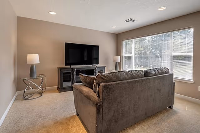 A cozy living room with a large gray sofa facing a flat-screen TV on a dark wooden TV stand. There are two modern table lamps on either side of the TV stand, one on a round glass side table and the other on the floor. A large window with blinds allows natural light to fill the room, which has beige walls and carpeted flooring.