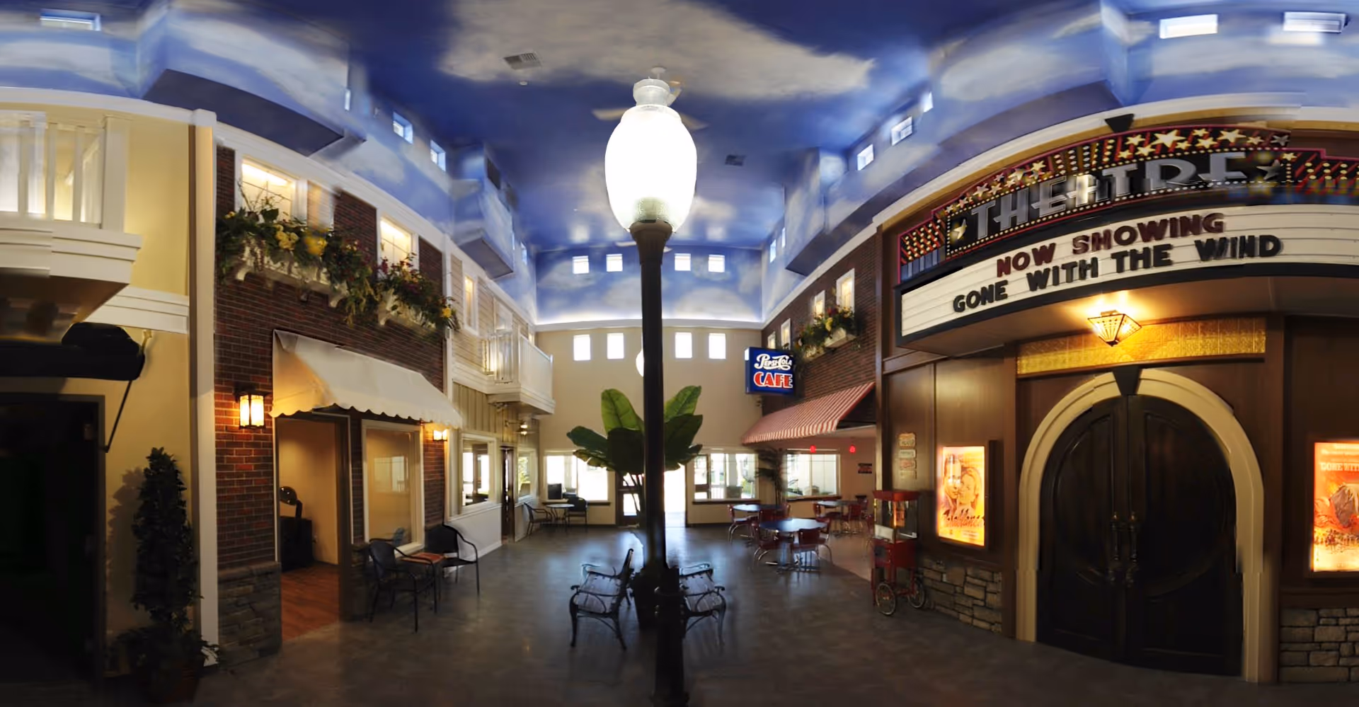 Interior of a senior living facility designed to resemble a small town street with a theater marquee showing 'Gone With The Wind', a Pepsi Cafe sign, benches, tables and chairs, and a lamppost in the center. The ceiling is painted with a blue sky and clouds, and there are decorative flower boxes on the walls.