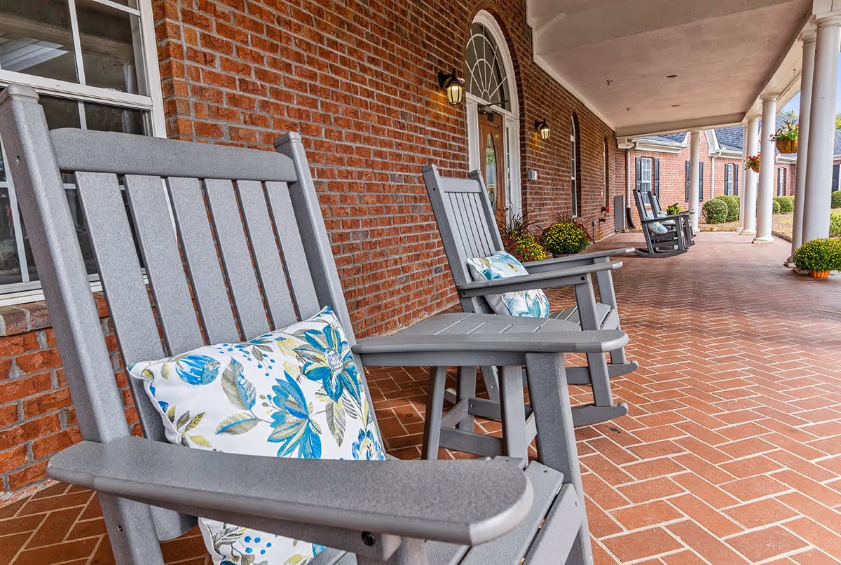 A covered outdoor porch area with gray rocking chairs featuring floral cushions, brick walls, white columns, and potted plants along the walkway.