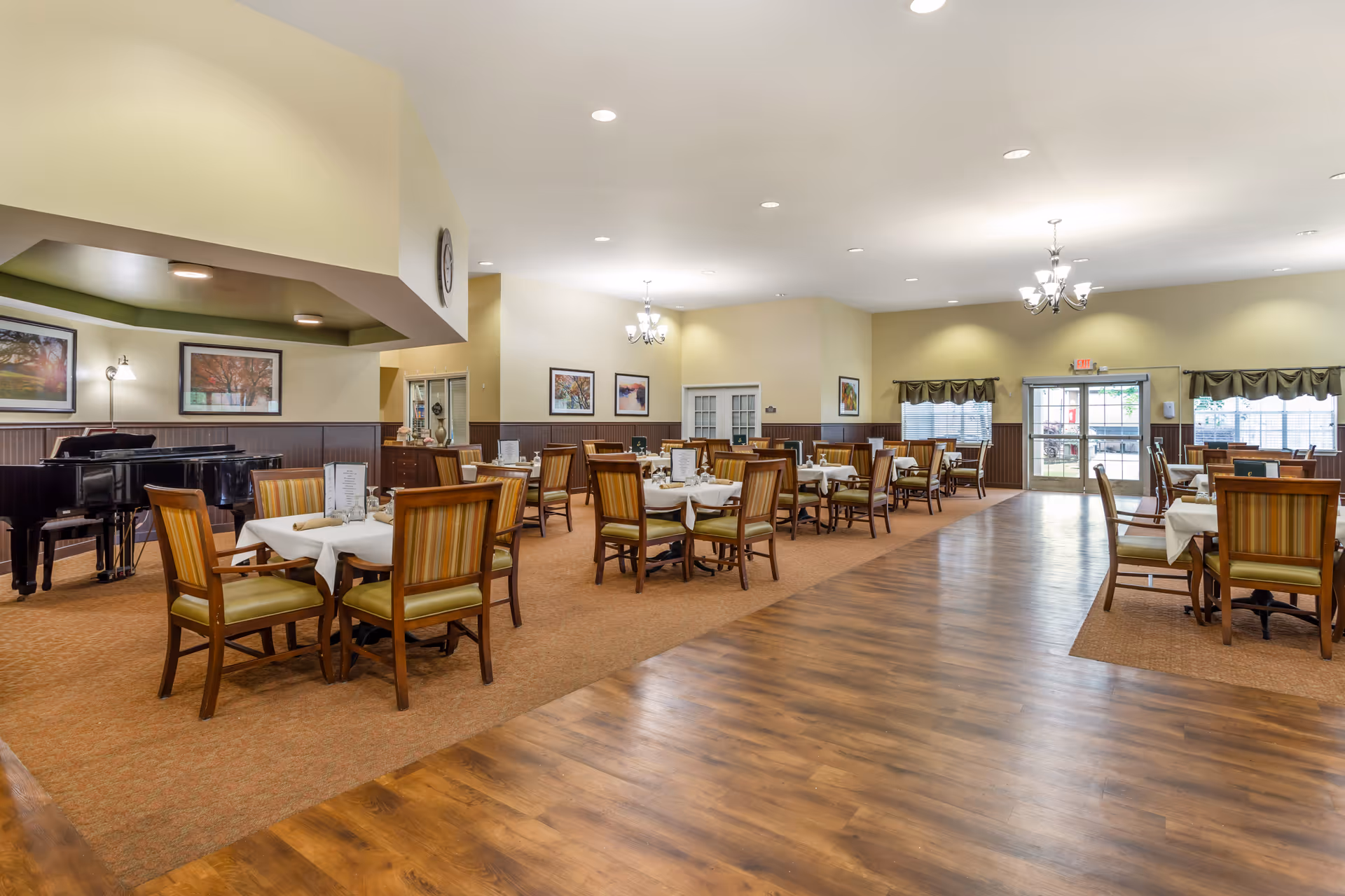 A spacious dining room with multiple tables covered with white tablecloths and set with dining ware. The room features wooden chairs with striped cushions, a polished wood floor pathway, carpeted areas, framed artwork on the walls, and a grand piano in the corner. Large windows and glass doors allow natural light to enter the room.
