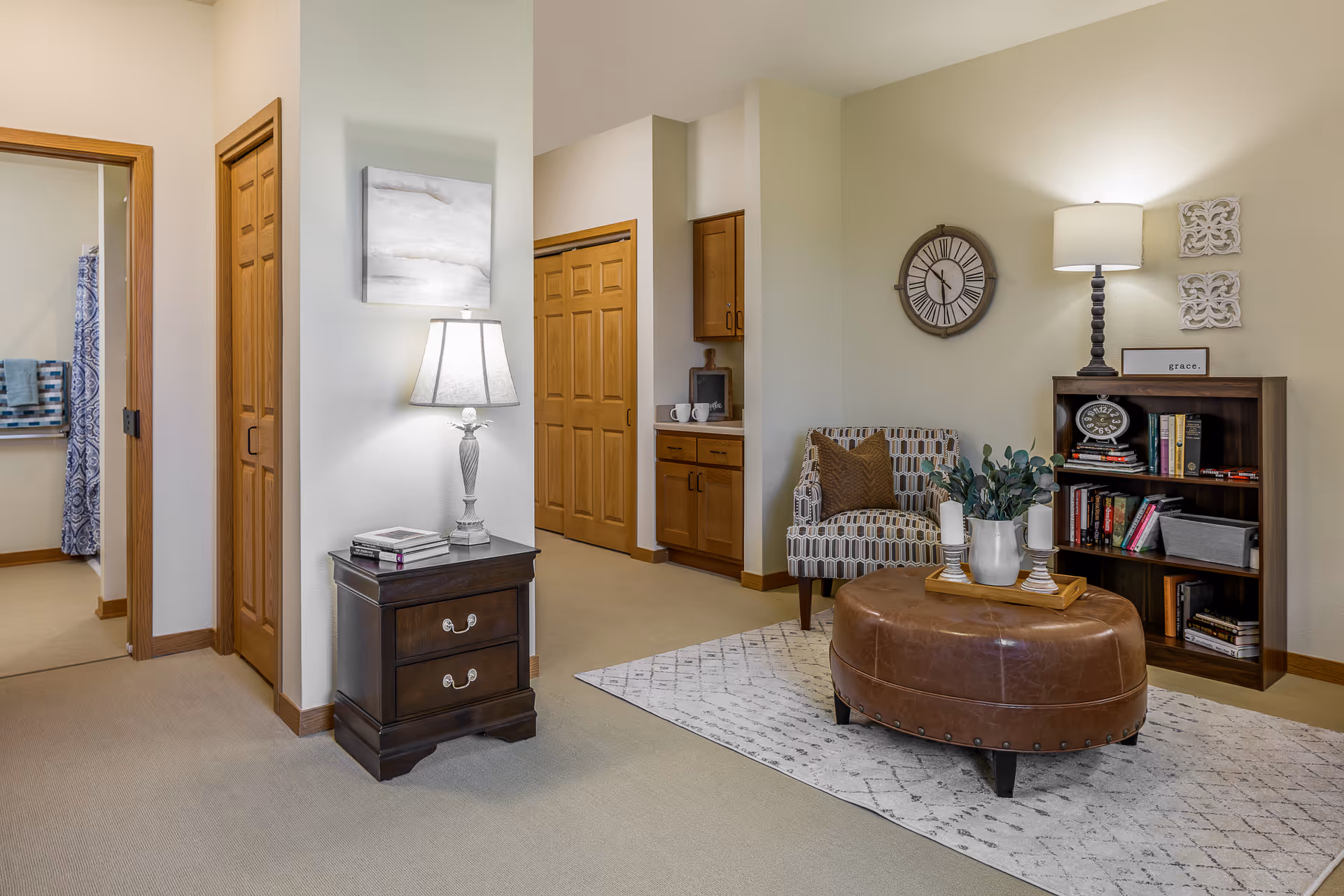 Well-lit living area with an upholstered chair, leather ottoman, side table and lamp, and a bookshelf against a neutral wall.