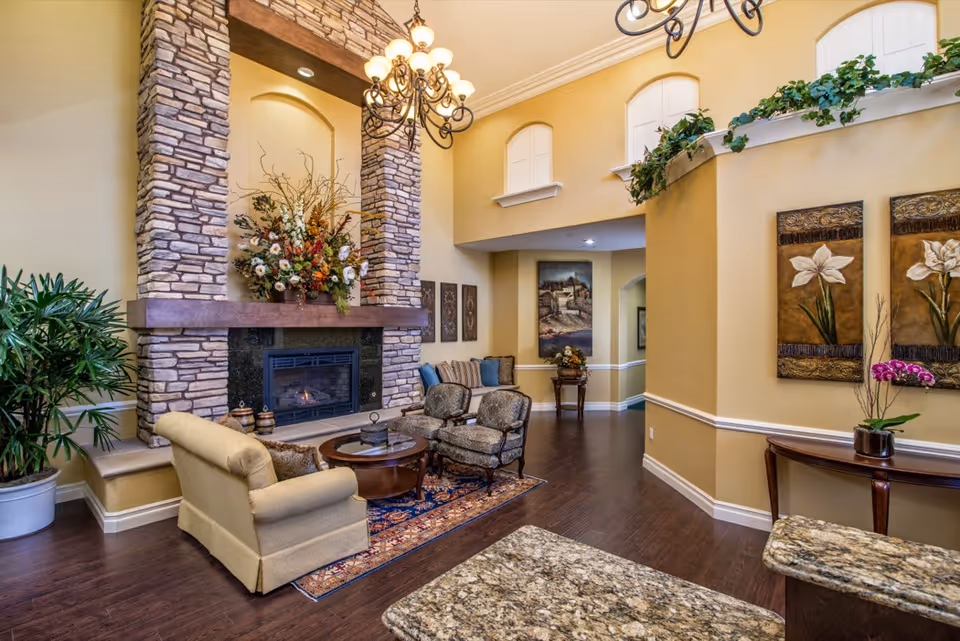 A cozy living room area with a tall stone fireplace decorated with a large floral arrangement. The room features a beige armchair, two patterned armchairs, a round wooden coffee table on a patterned rug, and hardwood floors. There are decorative paintings on the walls, potted plants, and a chandelier hanging from the ceiling. The walls are painted a warm yellow color with white trim.