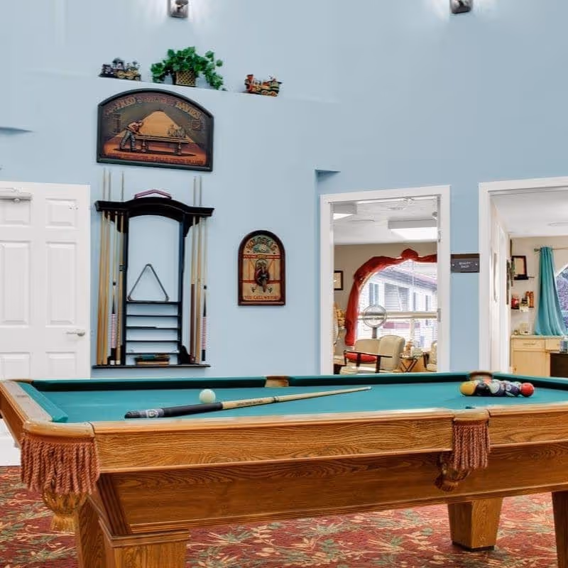 A recreational room with a wooden pool table featuring a green felt surface, pool cues, and billiard balls. The walls are painted light blue and decorated with a pool-themed sign and a rack holding pool cues. Two open doorways reveal adjacent rooms with seating and kitchen areas.