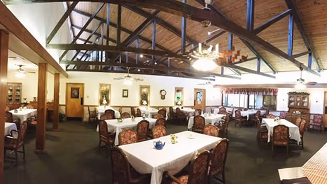 A spacious dining room with multiple tables covered in white tablecloths and surrounded by upholstered chairs. The room features a high wooden ceiling with exposed beams and ceiling fans. The walls are decorated with framed pictures and clocks, and there are large windows with curtains allowing natural light to enter.