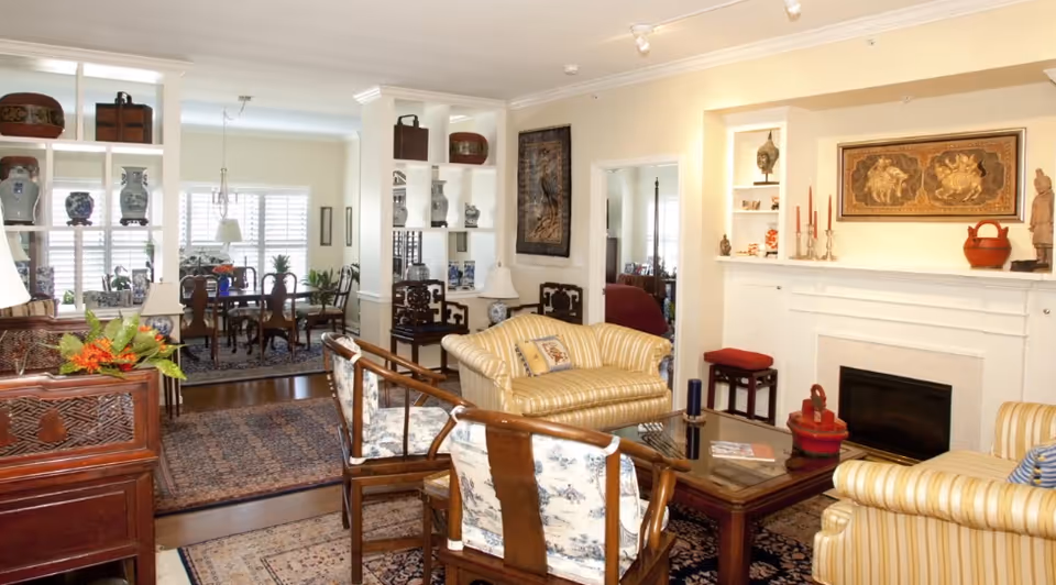 A cozy living room with striped yellow sofas, wooden chairs with floral cushions, a wooden coffee table, and a white fireplace adorned with decorative items and artwork. In the background, a dining area with a wooden dining table and chairs is visible, separated by a white shelving unit displaying vases and decorative objects.
