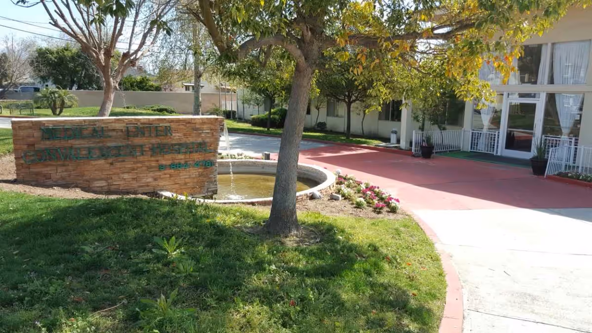 Outdoor view of the Medical Center Convalescent Hospital entrance area with a stone sign, a small fountain, trees, green grass, and a pathway leading to the building entrance with large windows and white curtains.