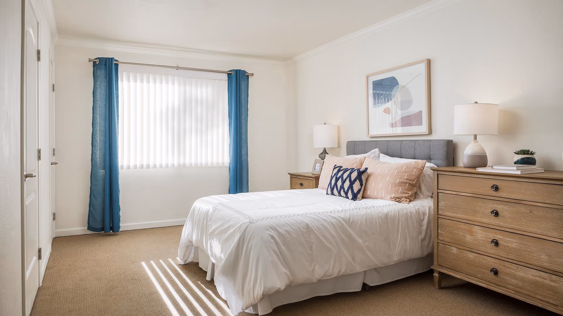 A bright bedroom with a neatly made bed featuring white bedding and decorative pillows. The room has beige carpeting, a window with vertical blinds and blue curtains, two wooden nightstands with lamps, a wooden dresser, and a framed abstract artwork on the wall above the bed.