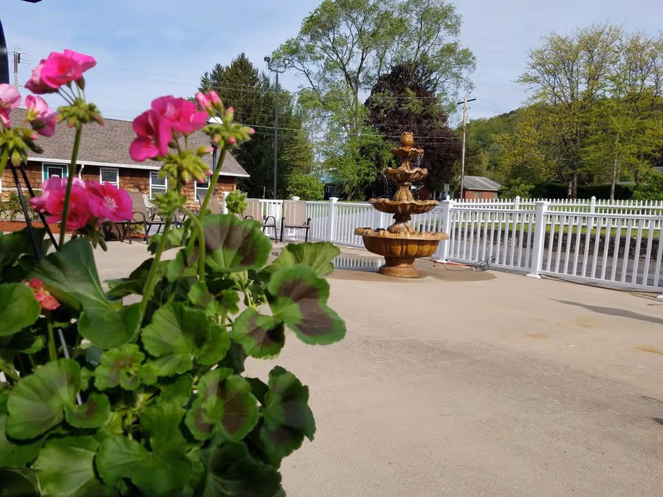 Outdoor patio area with a multi-tiered water fountain, white picket fence, several chairs, and a building with brown siding in the background. Pink flowers and green leaves are in the foreground.