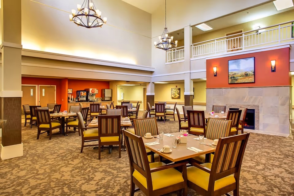 A spacious dining room with multiple wooden tables and chairs arranged neatly. Each table is set with cups, glasses, napkins, and small flower centerpieces. The room features high ceilings with two large chandeliers, a fireplace with a painting above it, and a beverage station in the back corner. The walls are painted in warm tones with a mix of beige and red accents.