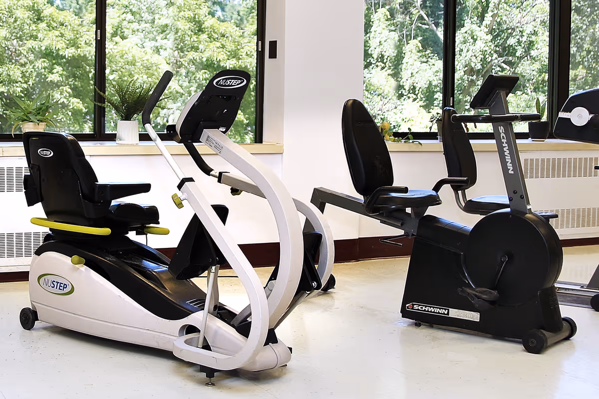 A bright exercise room with two recumbent stationary bikes positioned near large windows showing green trees outside. The bikes are black and white with comfortable seats and pedals, designed for seated cycling workouts.