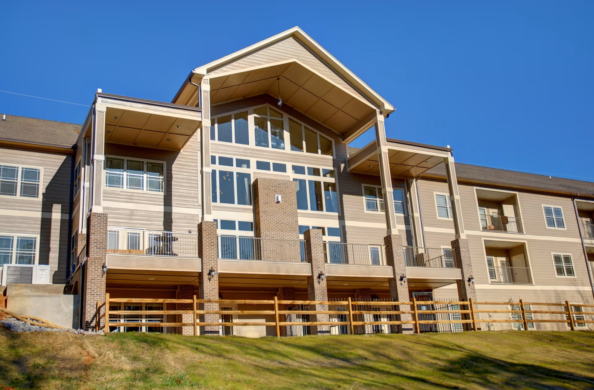 Exterior view of a multi-story residential building with large windows, balconies, and a sloped roof under a clear blue sky. The building has a combination of brick and siding materials, with a wooden fence and grassy area in the foreground.