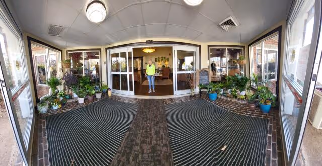 Entrance area of City Center Village Senior Community showing a covered entryway with large floor mats, potted plants on either side, glass doors leading inside, and a person standing just inside the doors.