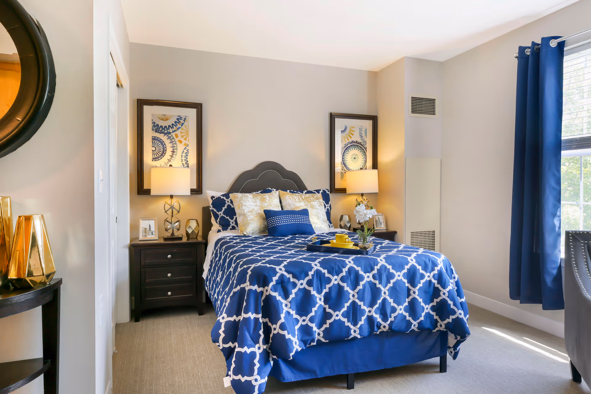 A neatly made bed with a blue and white patterned bedspread and multiple pillows in a well-lit bedroom. There are two nightstands on either side of the bed, each with a lamp and decorative items. Two framed artworks hang above the nightstands. A window with blue curtains allows natural light into the room. A round mirror and gold decorative vases are visible on a small table to the left.