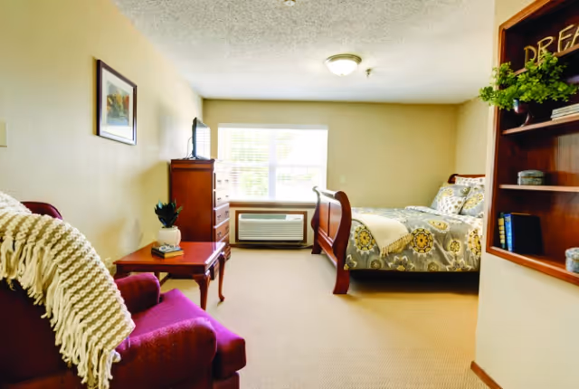 A cozy bedroom in Oak Hill Place featuring a wooden bed with patterned bedding, a wooden dresser with a TV on top, a window with blinds, a red armchair with a white knitted throw, a small wooden side table with a plant and books, and a built-in wooden shelf with decorative items.