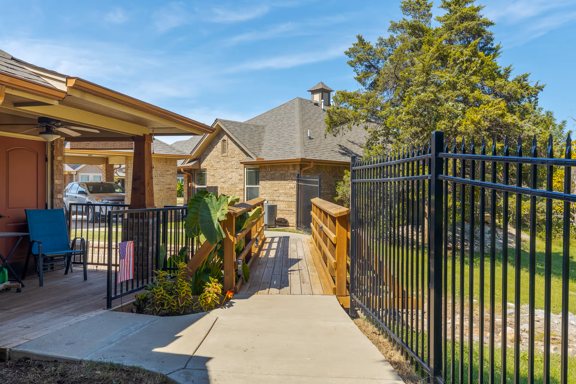 Wooden ramp and covered patio leading to a brick building, flanked by a black metal fence and trees under a blue sky.