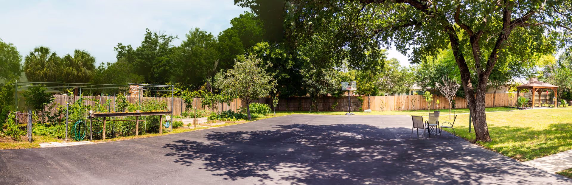 Outdoor area with a paved basketball court, a basketball hoop, a small table with two chairs under a tree, a garden area with plants and a hose, and a wooden gazebo in the background surrounded by trees and a wooden fence.