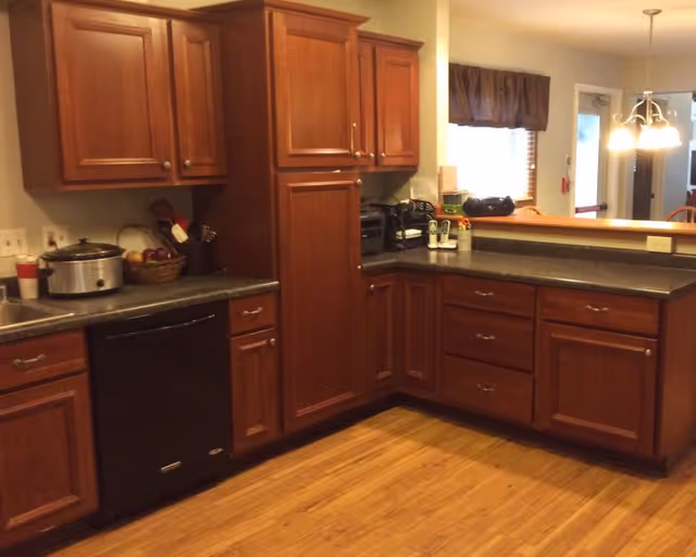 Kitchen with cherry wood cabinets, dark countertops, a black dishwasher, and hardwood floors opening to a dining area.