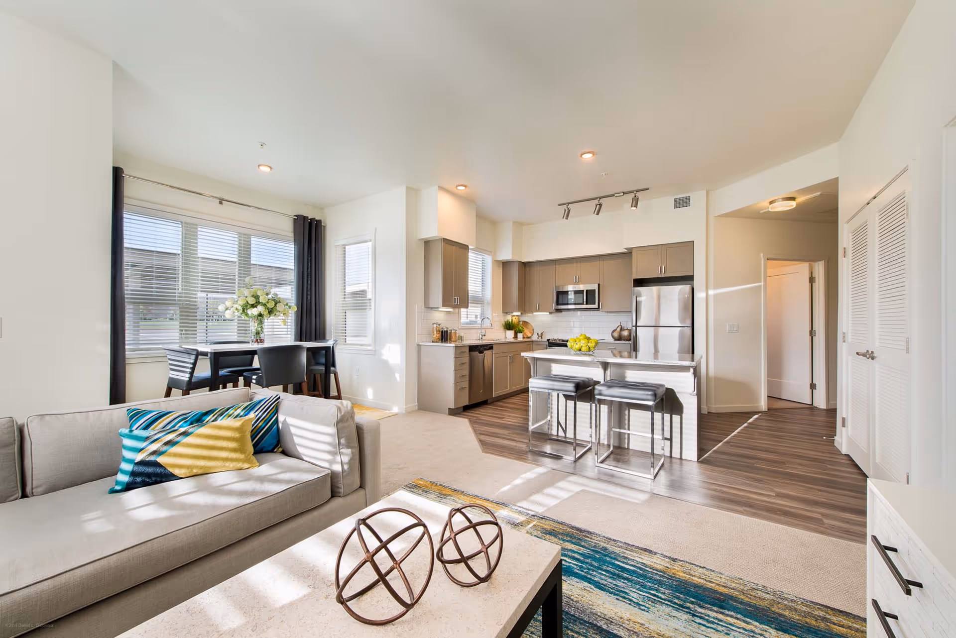 Bright open-plan living room and kitchen with a sofa, coffee table, dining table, and a kitchen island with bar stools.