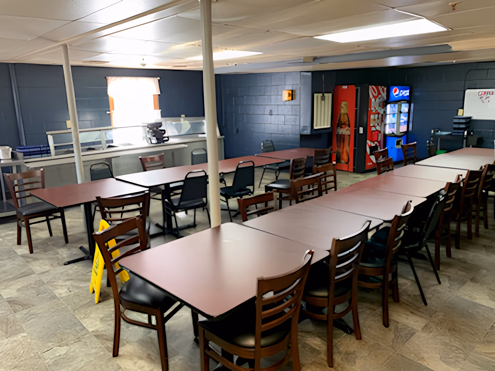 Large dining room with rows of tables and chairs, vending machines along the back wall, and a service counter in the background.