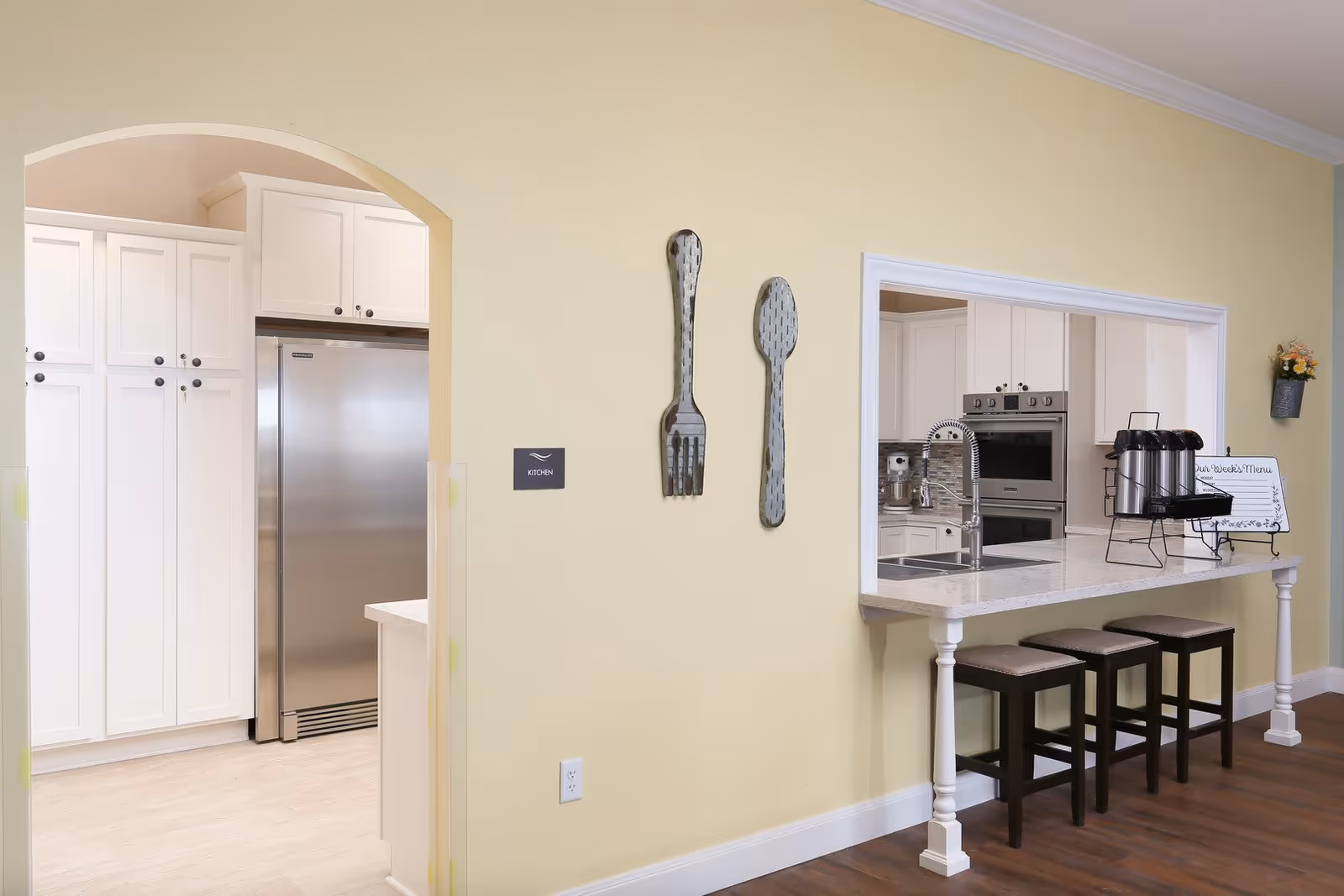 Interior view of a kitchen area in Serenity Oaks Assisted Living and Memory Care featuring white cabinetry, a stainless steel refrigerator, a countertop with three stools, a sink with a modern faucet, and decorative oversized fork and spoon wall hangings on a light yellow wall.