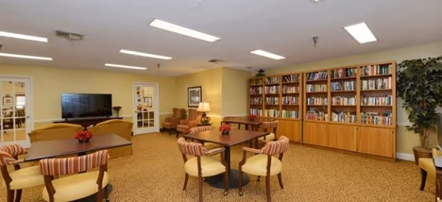 Well-lit senior lounge with round tables and chairs, a TV, bookshelves, and additional seating.