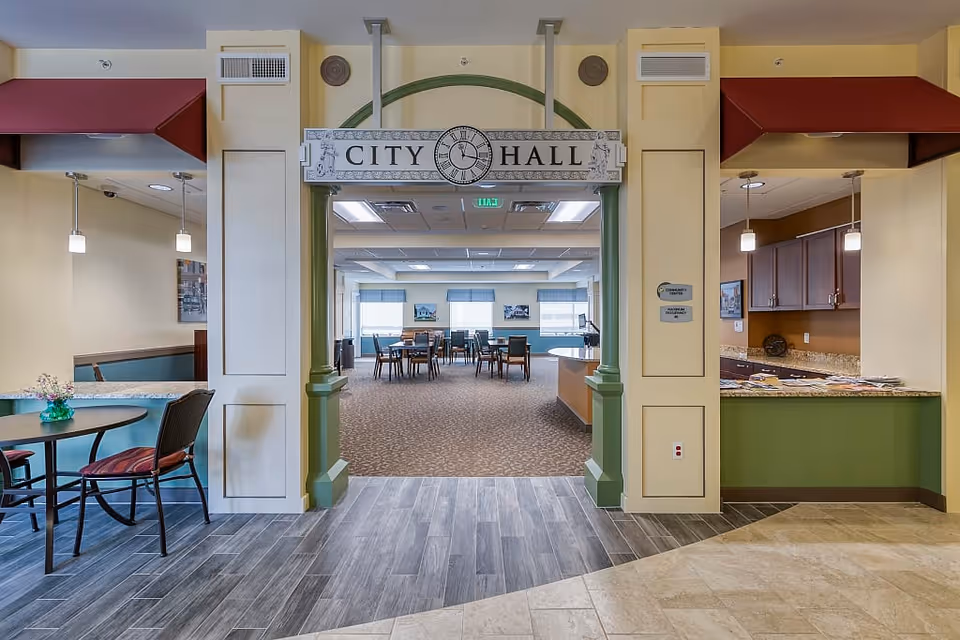 Interior view of a senior living facility area named City Hall, featuring a wide entrance with green pillars and a sign overhead. Inside, there are tables and chairs arranged in a spacious room with large windows letting in natural light. On the left, there is a small round table with chairs and a vase with flowers, and on the right, a counter with cabinets and pendant lights above.