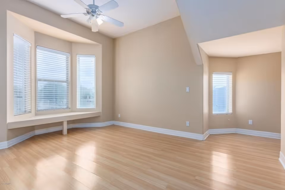 Empty room with light wood flooring, beige walls, a ceiling fan with light, and large bay windows with white blinds. There is a small alcove area with a window and white blinds.