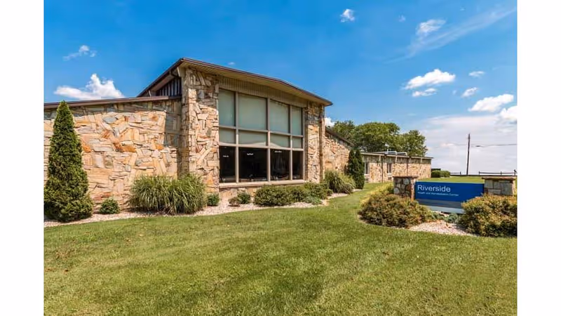 Exterior view of a single-story stone building with large windows, surrounded by green grass, shrubs, and small trees under a blue sky with scattered clouds. A sign in front reads 'Riverside Health and Rehabilitation Center.'