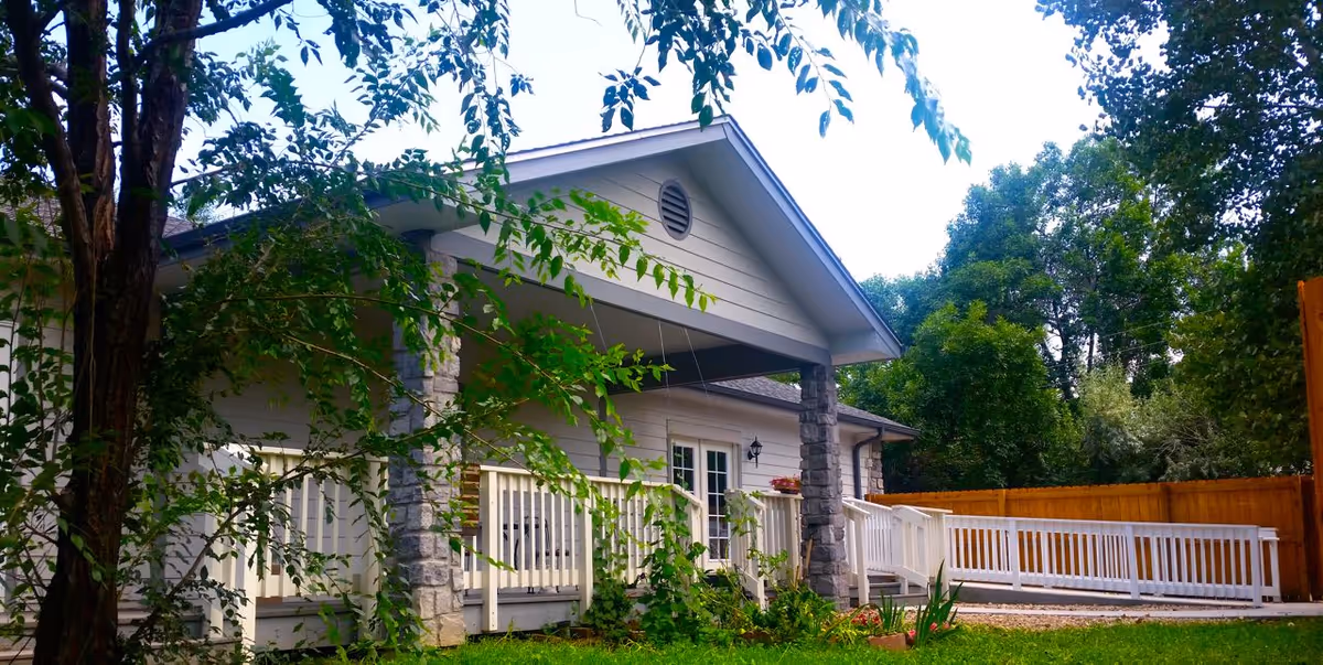 Exterior view of a single-story building with a covered porch supported by stone pillars, surrounded by trees and greenery. The porch has white railings and a wheelchair-accessible ramp leading to the entrance.