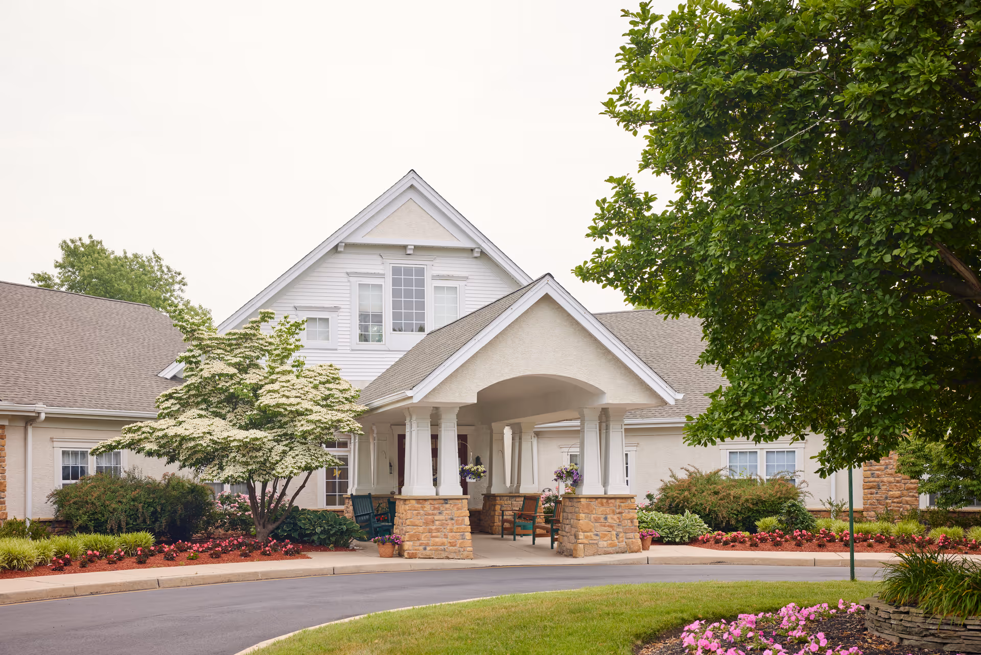 Front exterior view of a senior living facility with a covered entrance supported by white columns and stone bases. The building has a beige and white facade with multiple windows, surrounded by well-maintained landscaping including trees, bushes, and flower beds.