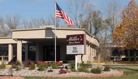 Front view of Miller's Merry Manor facility with flag, sign, and landscaped entrance.