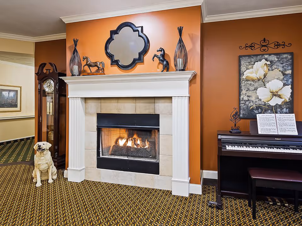 Cozy interior room with a lit fireplace surrounded by beige tiles and a white mantel. On the mantel are decorative items including two horse figurines and two tall vases. To the left of the fireplace is a tall grandfather clock and a statue of a sitting dog. To the right is a piano with sheet music and a painting of white flowers on an orange wall. The floor is covered with a patterned carpet.