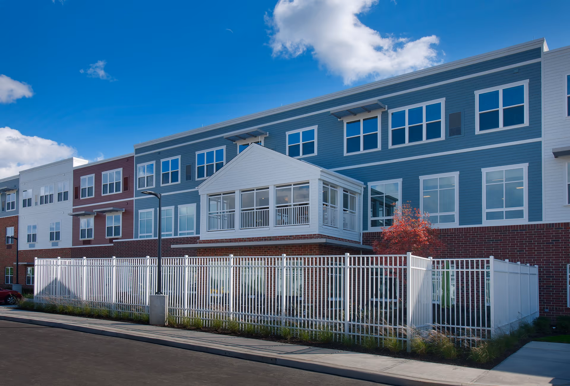Exterior view of a multi-story senior living facility building with a combination of brick and siding in blue, white, and red colors. The building has many windows and a white fenced area in front. The sky is clear with a few clouds.