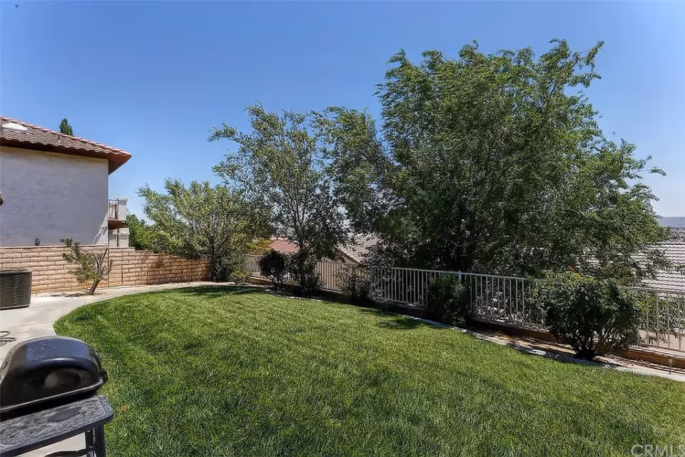A backyard area with a well-maintained green lawn, several trees and bushes along a metal fence, a concrete patio with a barbecue grill, and a clear blue sky overhead.