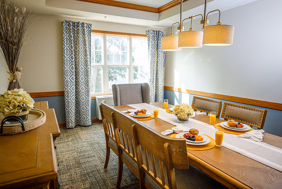 A bright dining room with a wooden table set for four people. The table has plates with muffins and fruit, glasses of orange juice, white napkins, and a white table runner. There are wooden chairs and a cushioned armchair around the table. The room has patterned blue and white curtains on the window, a sideboard with a flower arrangement, and a modern three-light pendant hanging above the table.