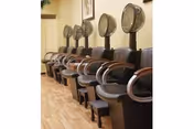 A row of vintage hair drying chairs with hooded hair dryers in a salon area featuring wooden flooring and framed artwork on the wall.