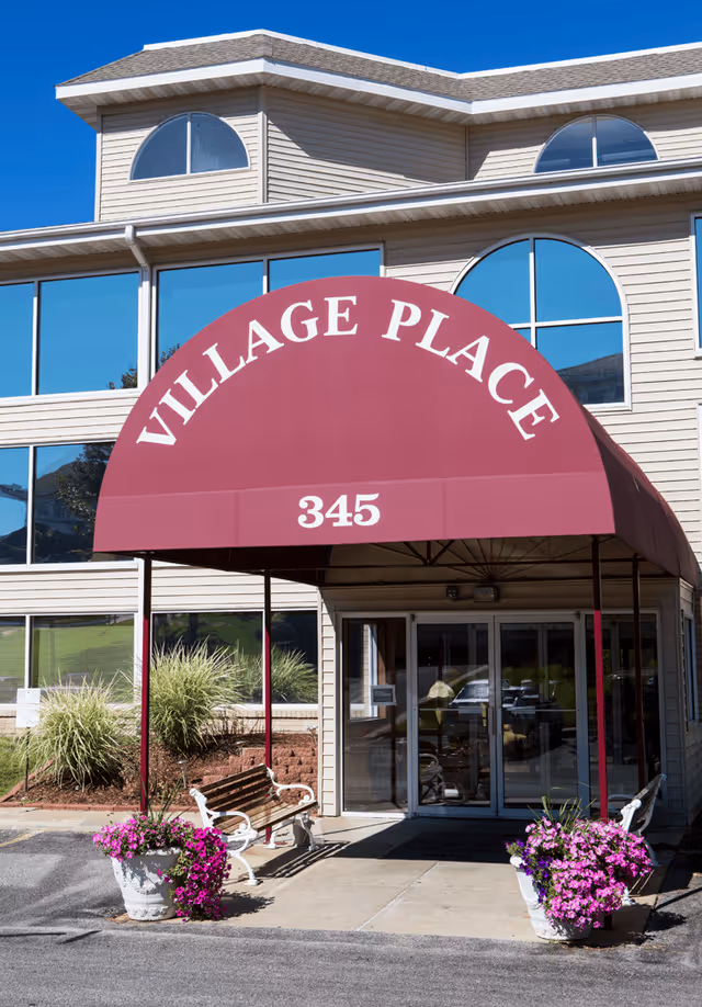 Entrance of the Village Place building with a maroon awning reading 'VILLAGE PLACE' and '345', flanked by benches and flower planters.