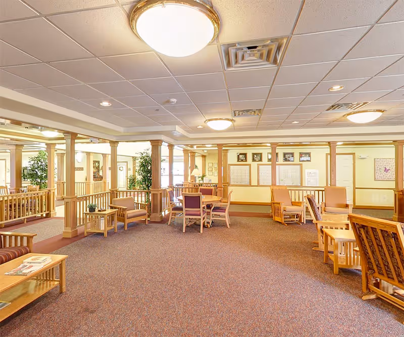 Spacious, well-lit senior living common area with multiple seating areas, tables, wooden columns and carpeted floor.