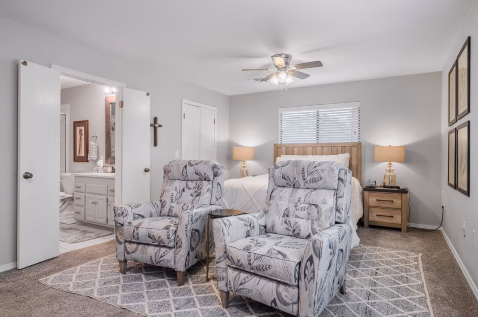 Well-lit bedroom with a bed, two patterned recliners in the foreground, nightstands, and an open door leading to a bathroom.