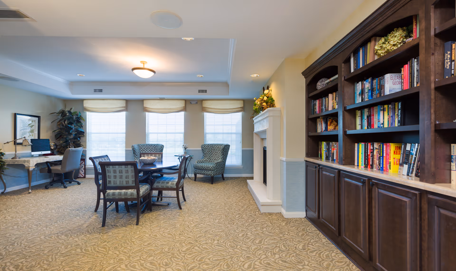 A cozy common area in an assisted living facility featuring a round table with four chairs in the center, two patterned armchairs near three large windows with blinds, a built-in dark wood bookshelf filled with books on the right, a white fireplace with floral decoration above it, and a desk with a computer and chair on the left side of the room.