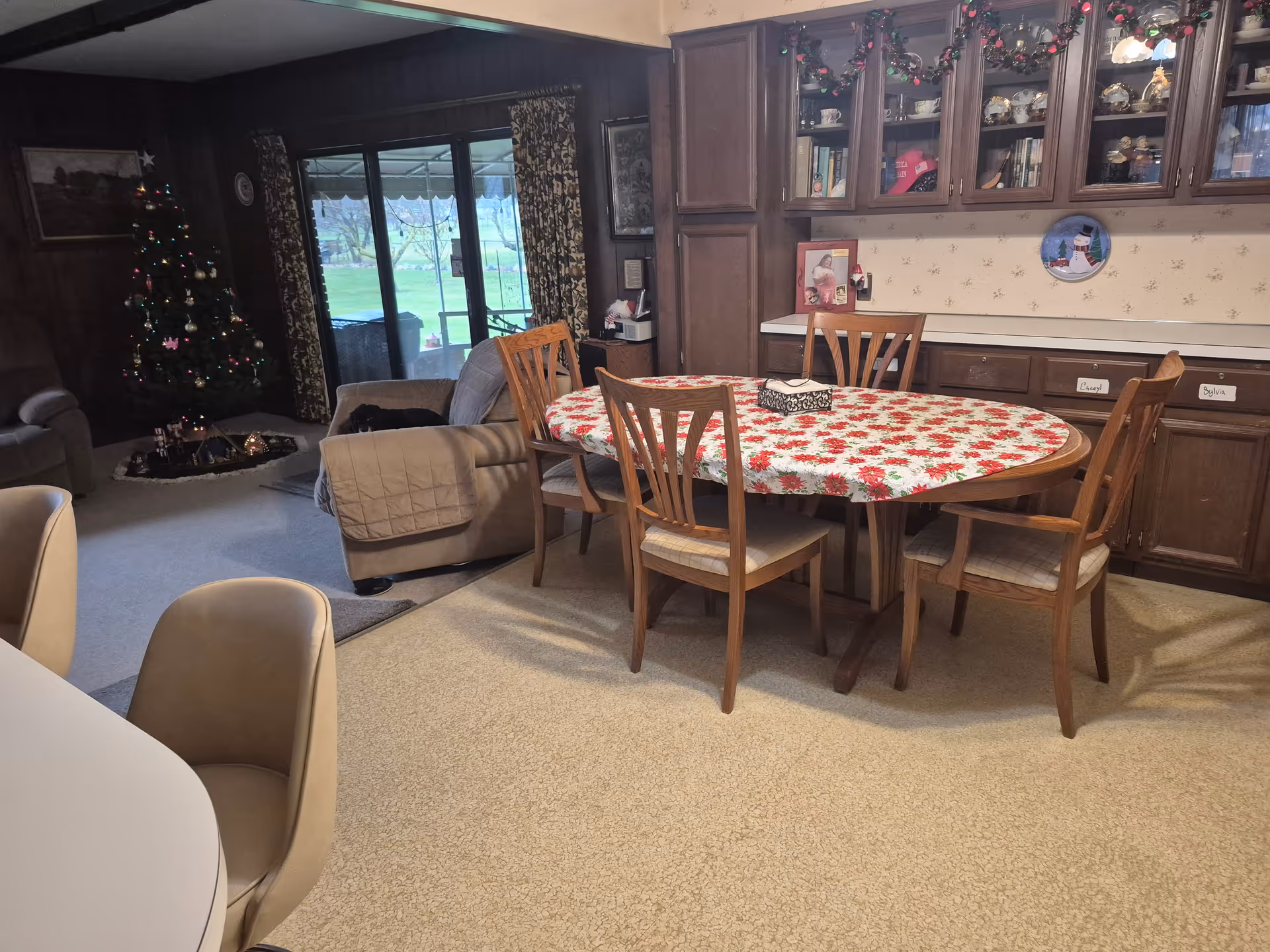 Interior view of a cozy living and dining area decorated for Christmas. A round dining table with a red and white floral tablecloth is surrounded by four wooden chairs. Behind the table, there are wooden cabinets with glass doors displaying various items and holiday decorations. To the left, a living room area features a Christmas tree with lights and ornaments, a recliner chair, and a sliding glass door leading outside. The room has carpeted floors and patterned curtains.
