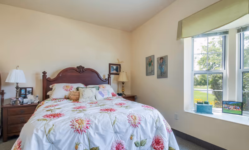 Sunlit bedroom with a wooden headboard, floral bedspread, nightstands with lamps, and a large window seat.