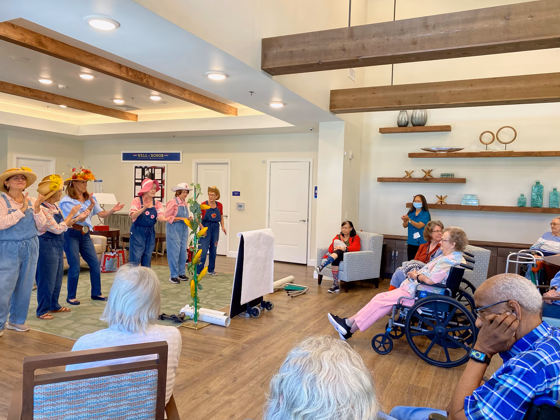 A group of elderly people seated in a common area watching a performance by six women dressed in overalls and colorful hats. The room has wooden beams on the ceiling, shelves with decorative items, and a sign on the wall that reads 'Wall of Honor'. Some audience members are seated in wheelchairs and others in chairs, with a staff member standing and clapping.