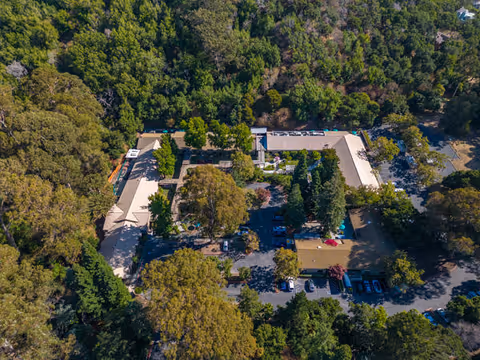 Aerial view of Silverado Belmont Hills Memory Care Community surrounded by dense green trees. The facility buildings are arranged in a U-shape with parking areas and pathways visible around the property.