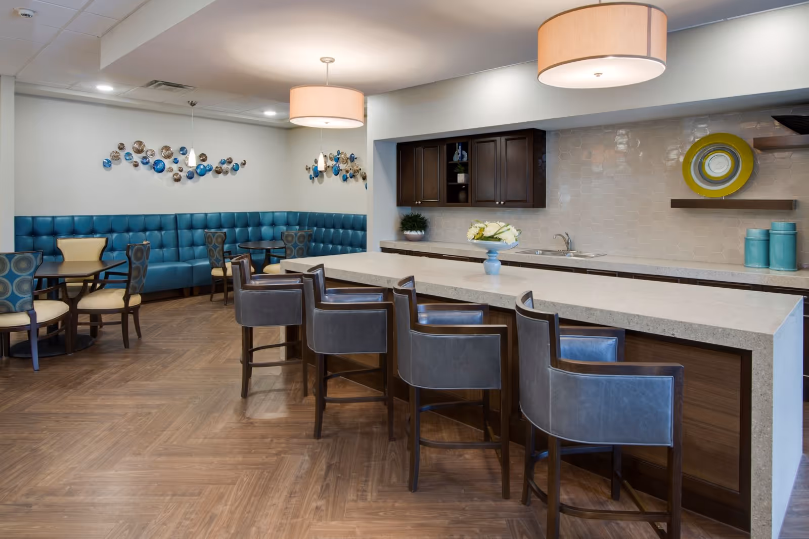 A modern dining area with a long kitchen island featuring five leather bar stools. The background includes a wall with dark wooden cabinets, a sink, and decorative plates on shelves. To the left, there are multiple small dining tables with chairs and a blue cushioned bench along the wall. The floor is wooden, and the ceiling has two large round pendant lights.