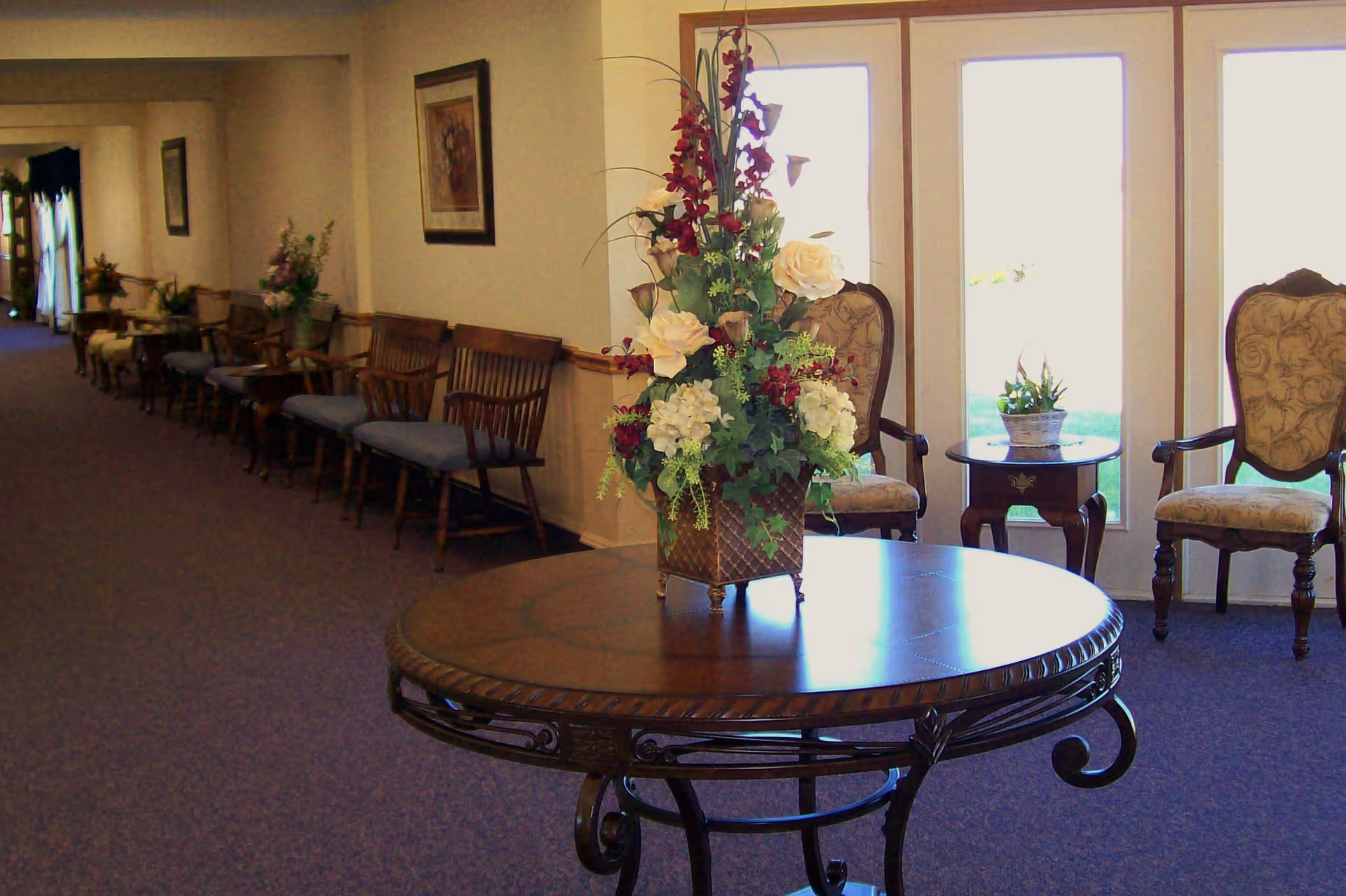 A seating area with a round wooden table topped with a floral arrangement, upholstered chairs, and a row of wooden benches along a carpeted corridor.