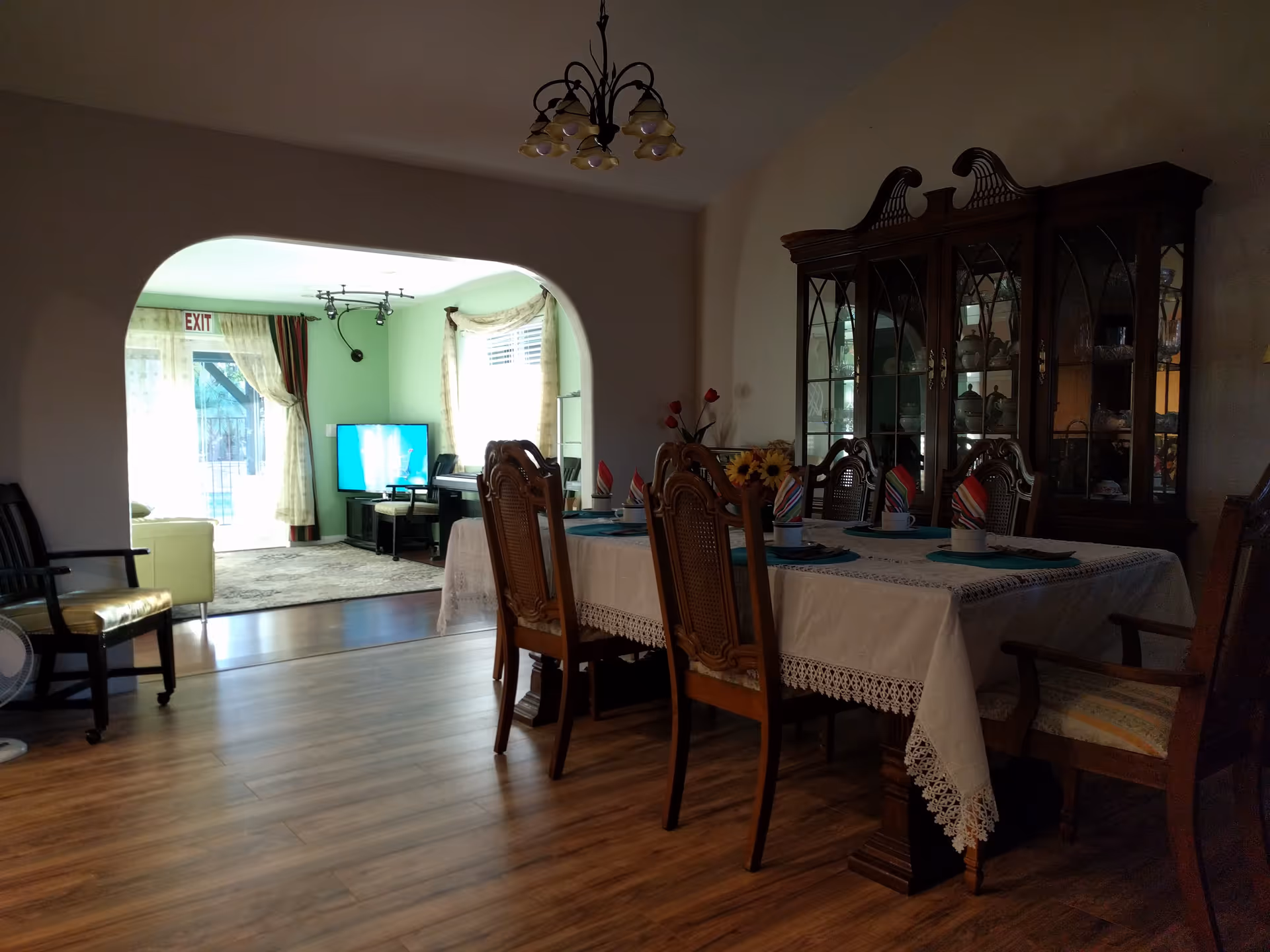 A dining room with a wooden table covered with a white lace tablecloth and set with cups, plates, and colorful napkins. There are six wooden chairs around the table. In the background, through an arched doorway, a living room area is visible with a green wall, a TV, a piano, and a glass door with curtains leading outside. The floor is wooden, and there is a wooden china cabinet against the right wall.