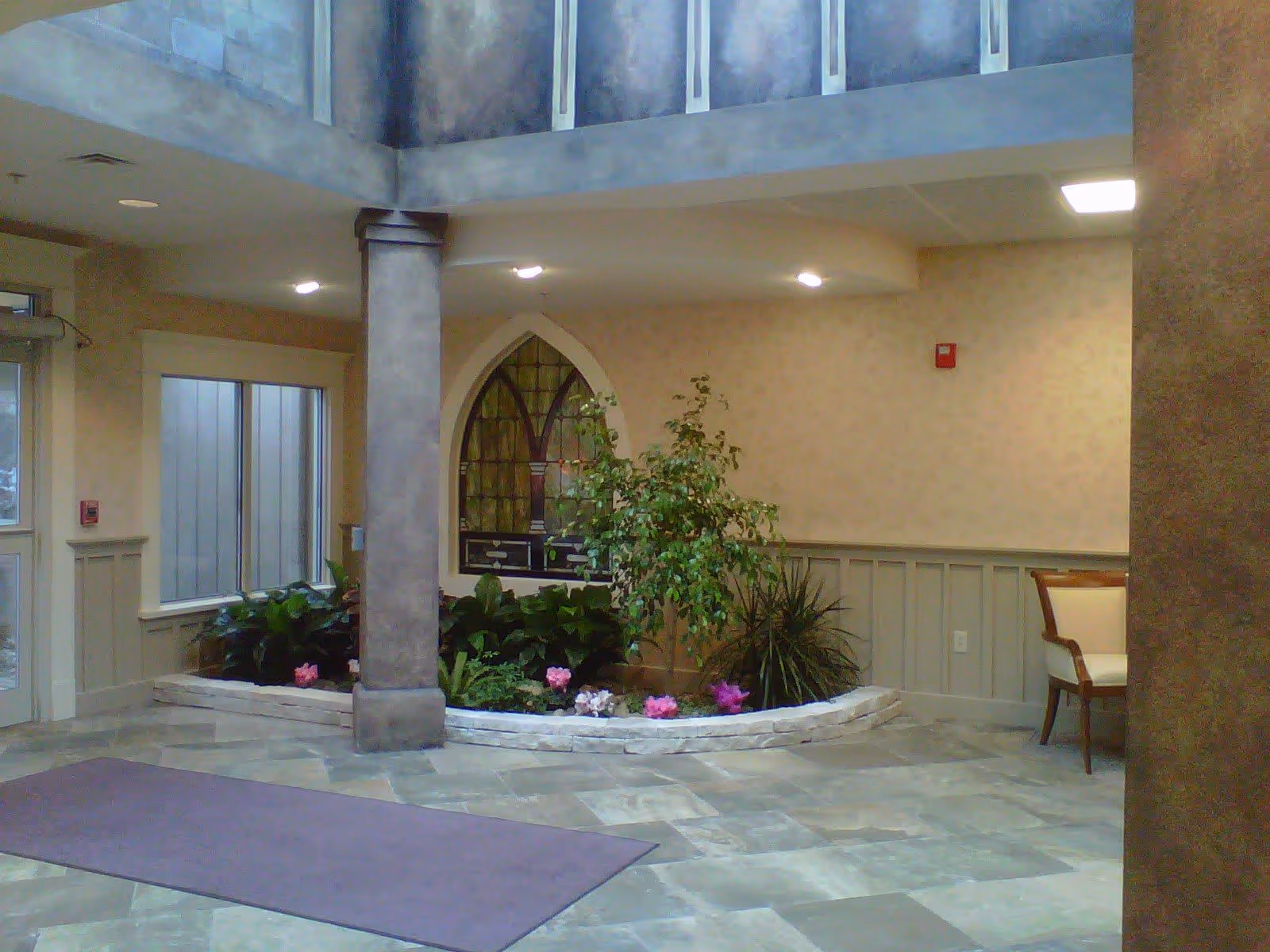 Indoor area with a stone-tiled floor, a purple rug, a central pillar, and a raised planter filled with green plants and pink flowers. The walls are beige with wainscoting, and there is a decorative arched window with stained glass. A wooden chair with a cream cushion is partially visible on the right side.