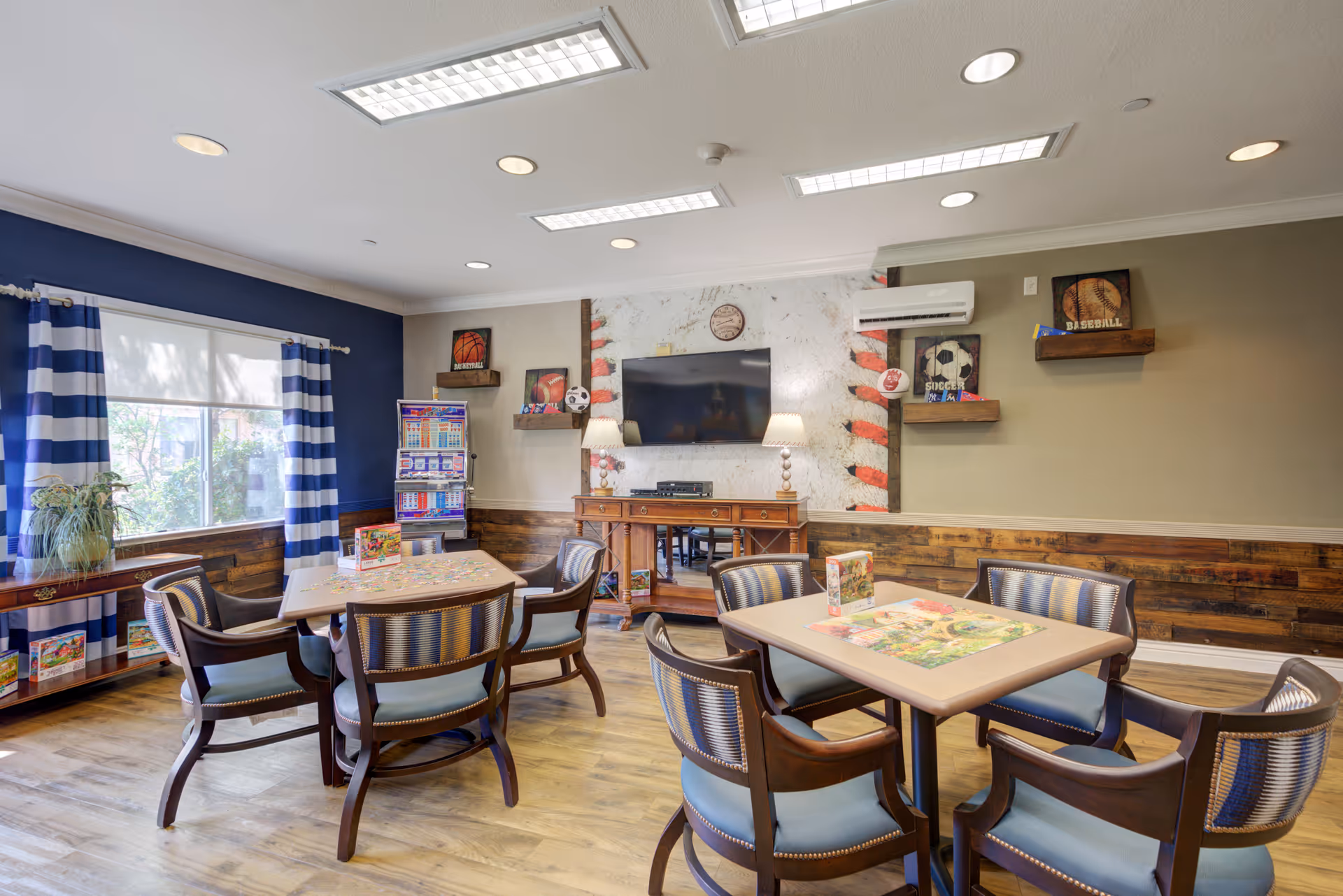 A bright and cozy activity room with two tables surrounded by wooden chairs with blue cushions. Each table has a partially completed jigsaw puzzle. The room features a large window with blue and white striped curtains, a wooden side table with a plant, and a wall decorated with sports-themed artwork and a large TV mounted above a wooden console table. The floor is wooden, and the ceiling has recessed lighting.